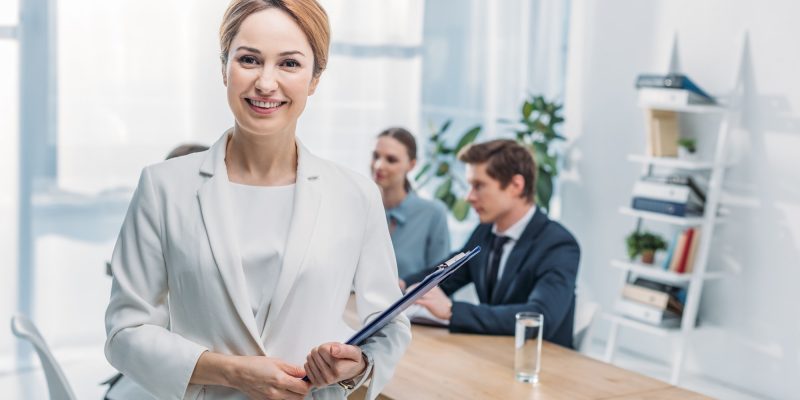 selective-focus-of-cheerful-recruiter-standing-with-clipboard-near-coworkers